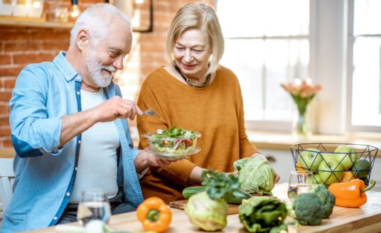 Cheerful senior couple eating salad standing together with healthy food on the kitchen at home.
