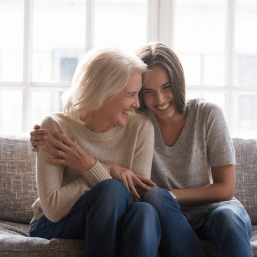 Two women sitting on a sofa, laughing together in front of large windows.