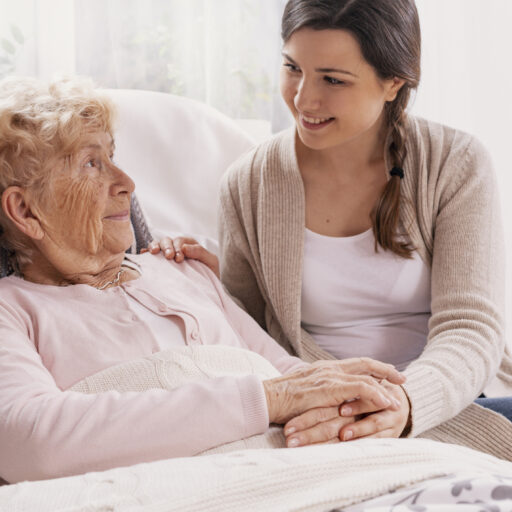 A young white woman sits next to her elderly female loved one in bed
