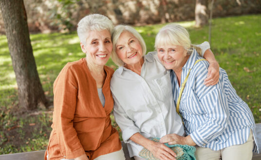 Three senior women embracing each other on a bench outside, maintaining their friendship for a healthy heart.