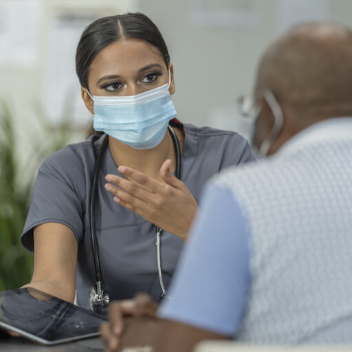 A senior patient and a female geriatric care manager meet in a medical clinic while wearing protective face masks to avoid the transfer of germs.