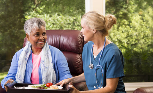 A senior woman speaks to her home health care nurse. As a senior ages, they may find themselves deciding between in-home senior care and assisted living. There are benefits to both, depending on the senior's needs.