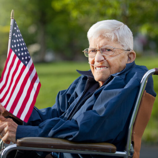 senior veteran sits in a wheelchair outside and holds an American flag