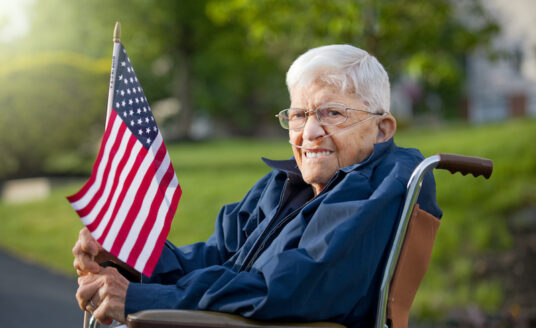 senior veteran sits in a wheelchair outside and holds an American flag