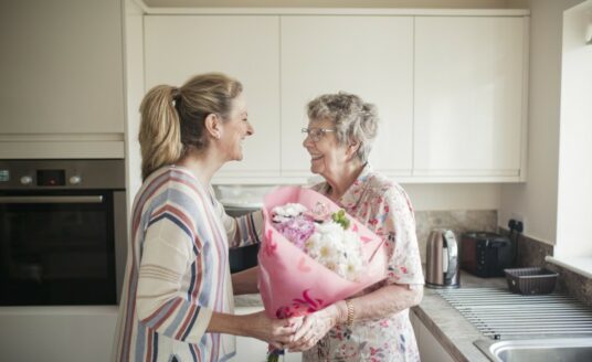 An older adult woman greets her senior mother, who is living with Alzheimer's, with a bouquet of flowers.